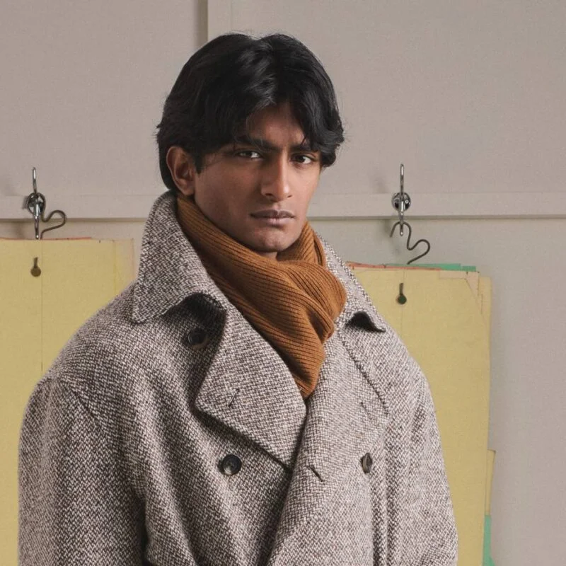 Person wearing a gray herringbone coat and orange scarf, posing in front of yellow file folders on a wall rack in an office-like setting.