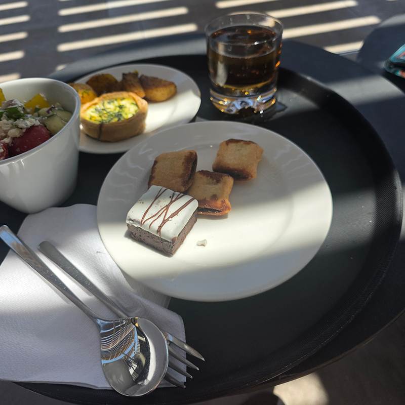 Outdoor table with a white plate of crostini and a frosted brownie, plus a bowl of salad and a glass of cola on a sunny patio.
