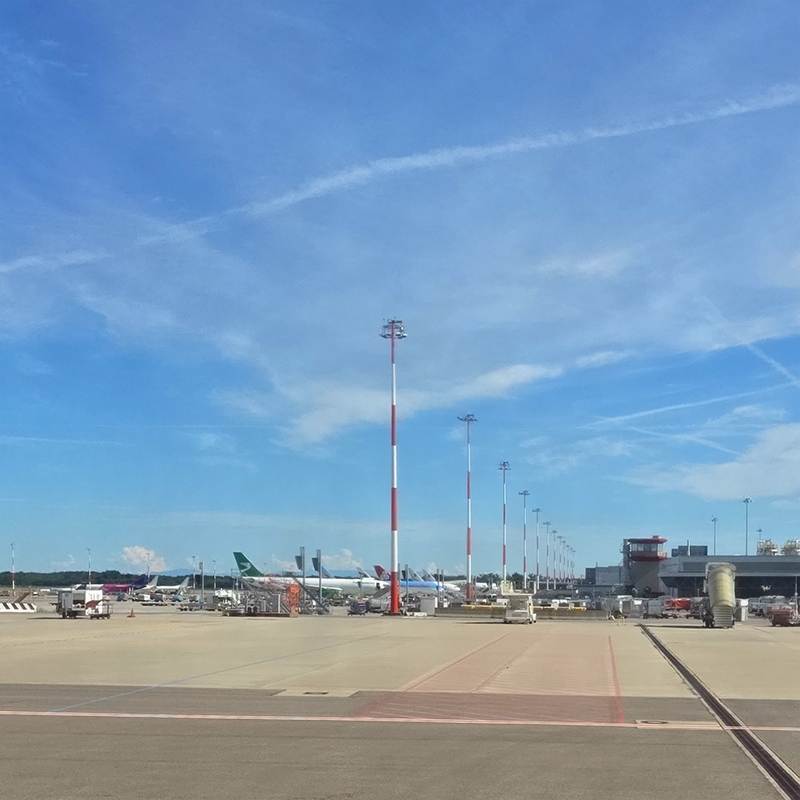 Airport gate area with several airplanes at the terminal, tall red-and-white light poles, and a blue sky.