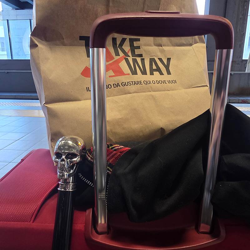 Red suitcase with a skull-knobbed cane leaning against it; a Take Away paper bag sits behind in a bright terminal area.