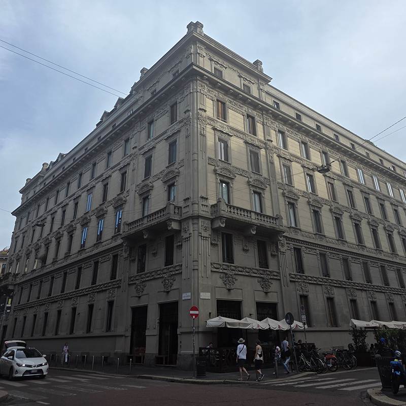 Corner historic building with ornate stone façade on a city street; pedestrians and a sidewalk café nearby.