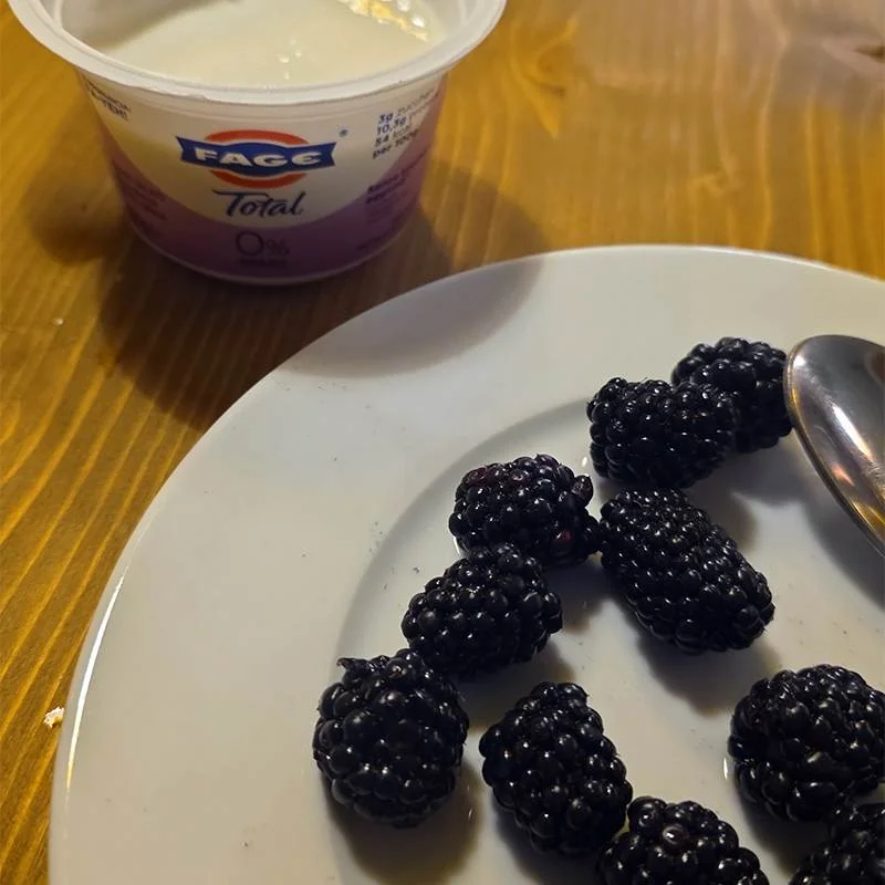 Plate of fresh blackberries with a spoon on a white dish; a cup of FAGE Total yogurt is visible in the background on a wooden table.