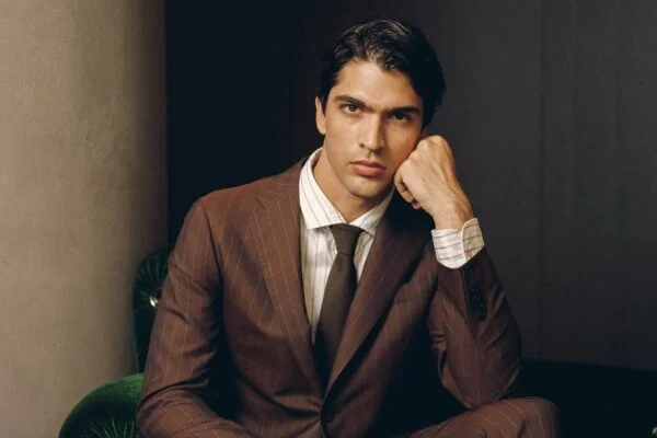 Portrait of a man in a brown pinstripe suit, seated on a green velvet chair, resting his chin on his hand and looking at the camera.