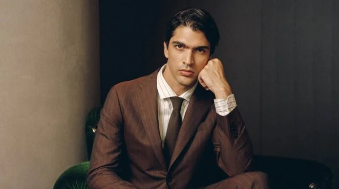 Portrait of a man in a brown pinstripe suit, seated on a green velvet chair, resting his chin on his hand and looking at the camera.