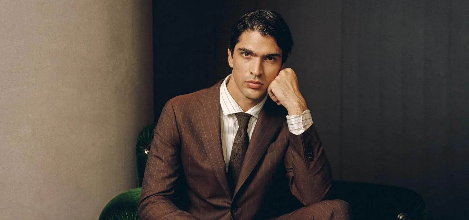 Portrait of a man in a brown pinstripe suit, seated on a green velvet chair, resting his chin on his hand and looking at the camera.