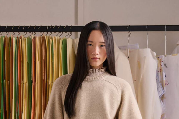 Woman with long black hair in a beige sweater stands in a fashion studio beside a rack of colorful folders and white garments behind her.