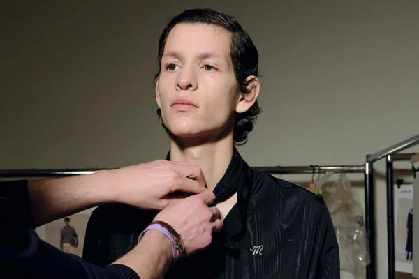Backstage scene of a stylist fastening the neck of a black shirt on a model, with clothing racks in the background.