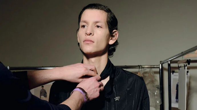 Backstage scene of a stylist fastening the neck of a black shirt on a model, with clothing racks in the background.