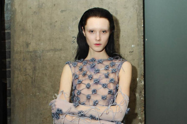 Model with pale makeup and dark hair wearing a sheer, blue-flowered mesh dress against a concrete wall.