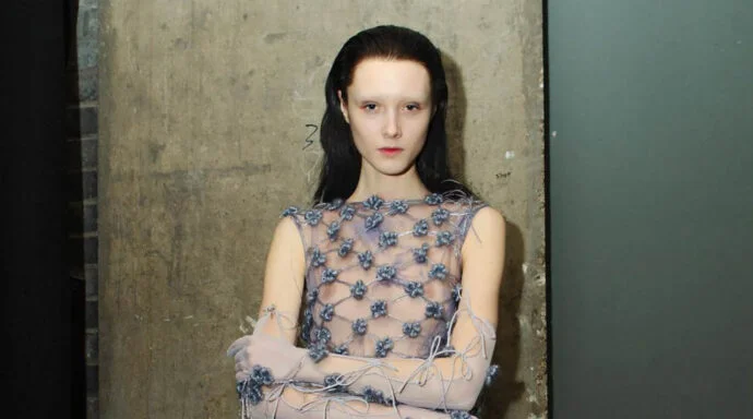 Model with pale makeup and dark hair wearing a sheer, blue-flowered mesh dress against a concrete wall.