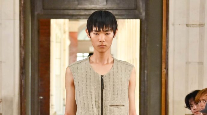Male fashion model walks runway wearing a sleeveless beige striped vest with a front zipper.
