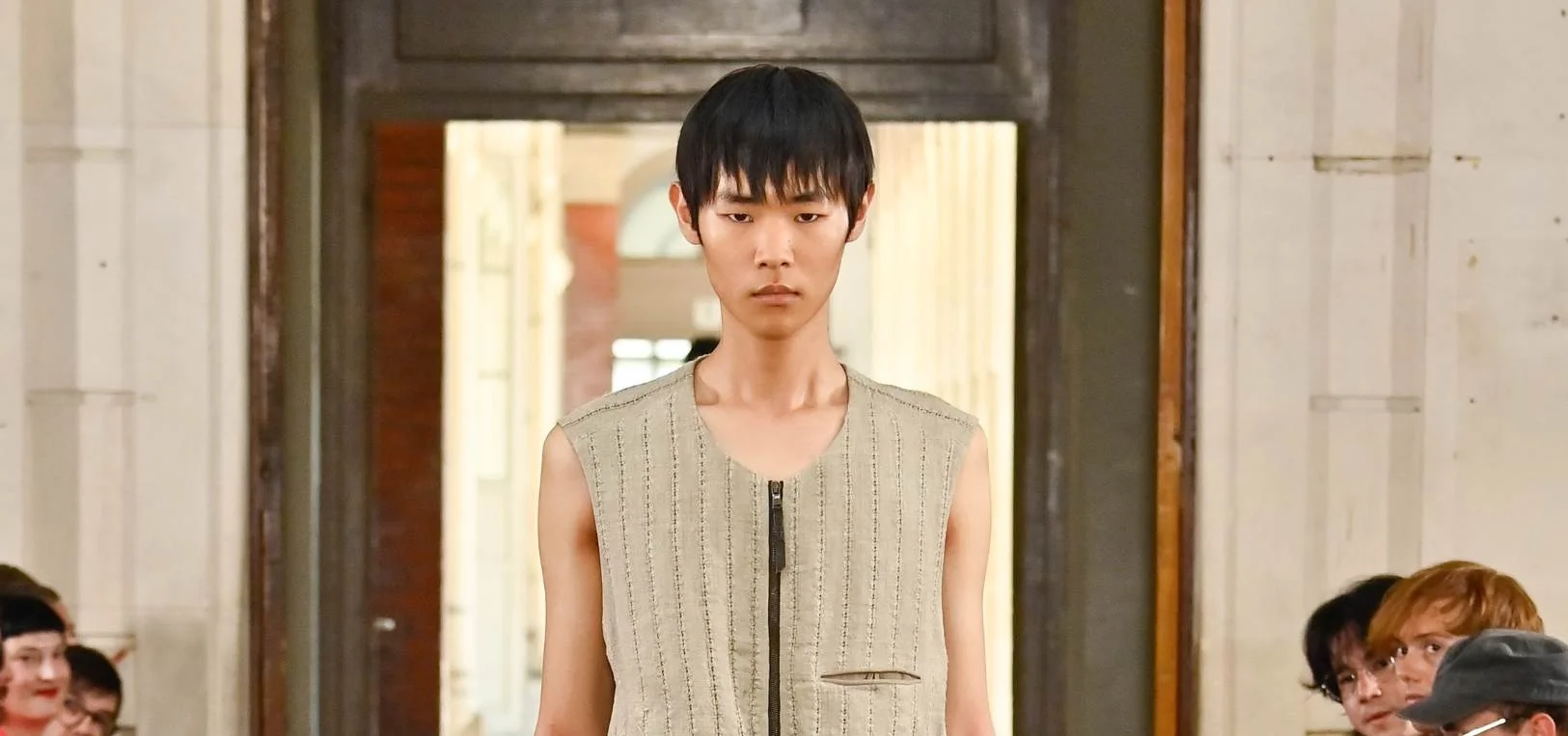 Male fashion model walks runway wearing a sleeveless beige striped vest with a front zipper.