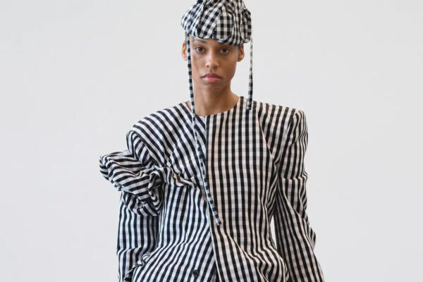 Model wearing a black-and-white gingham suit with oversized sleeves and a matching checkered cap, posed in a studio against a pale background.