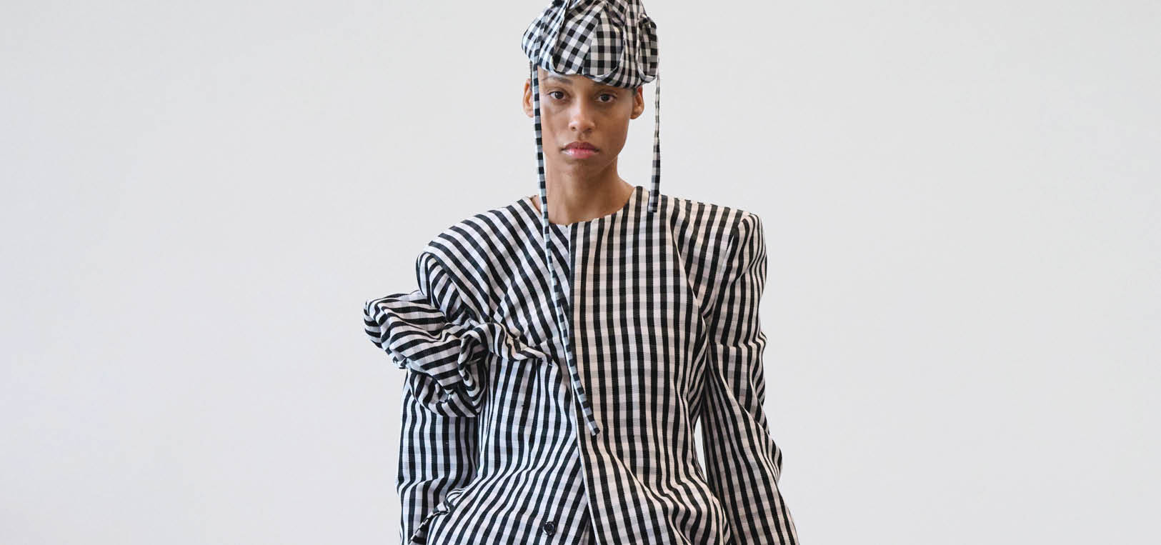 Model wearing a black-and-white gingham suit with oversized sleeves and a matching checkered cap, posed in a studio against a pale background.